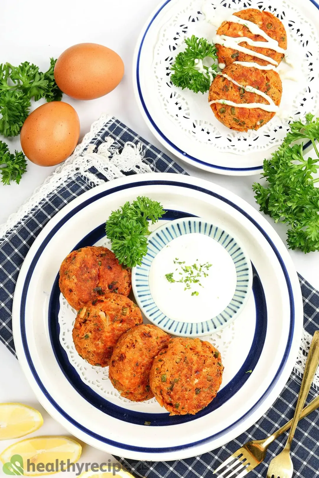 Two plates of fried salmon patties with the white yogurt sauce and some egg and parsley for decoration.