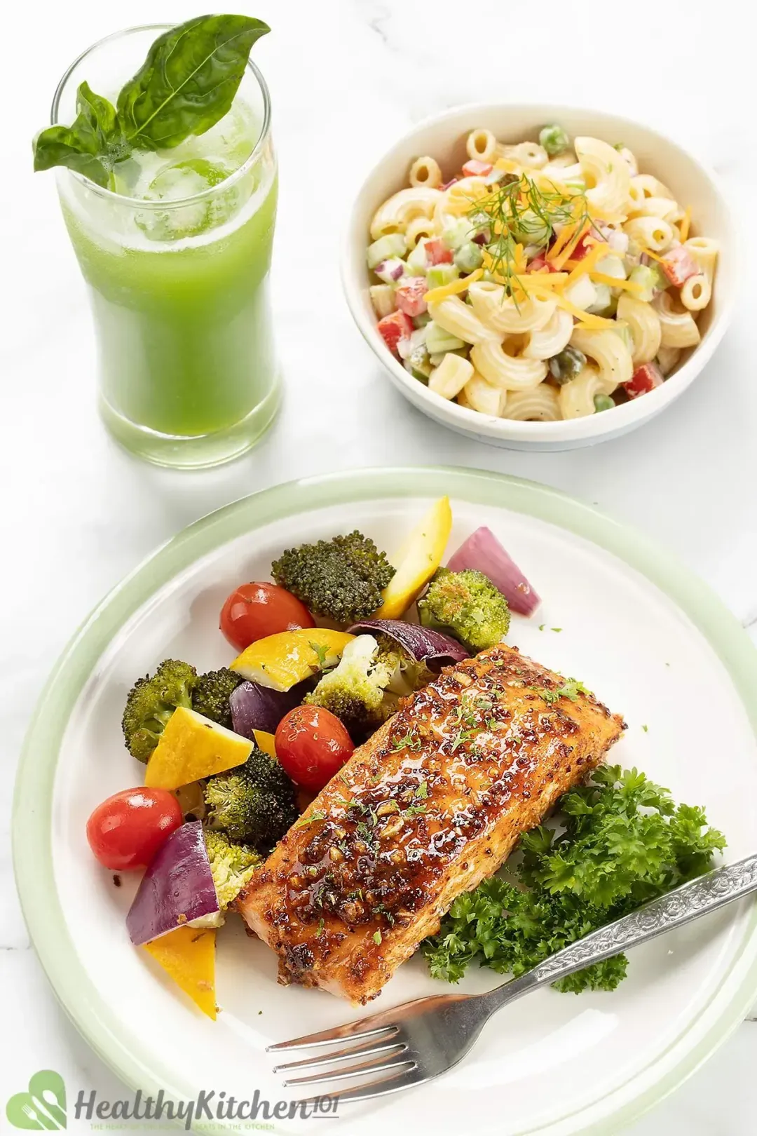 A plate containing a salmon fillet, parsley, cherry tomatoes, broccoli, and other veggies placed next to a small bowl of pasta salad and a glass of green juice