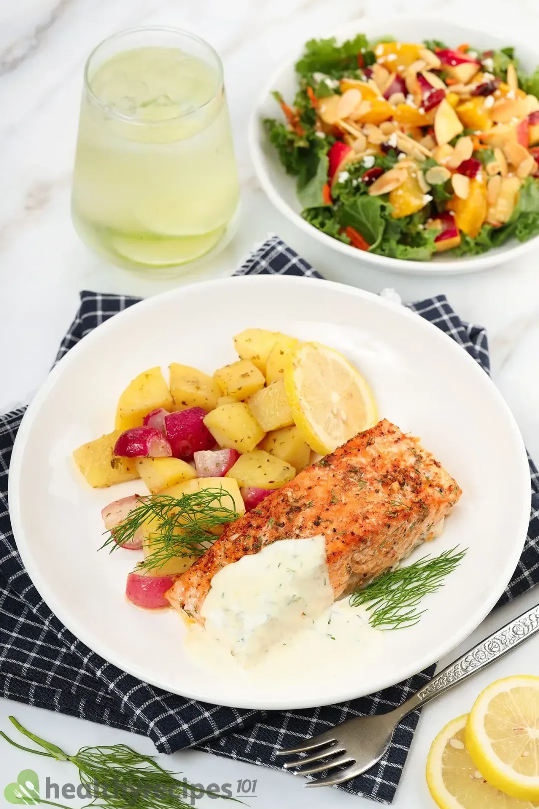 A of baked salmon, potato cubes, radish cubes, and fresh dill laid on top of a blue tablecloth and near a plate of salad, a glass of lemon juice, and a fork