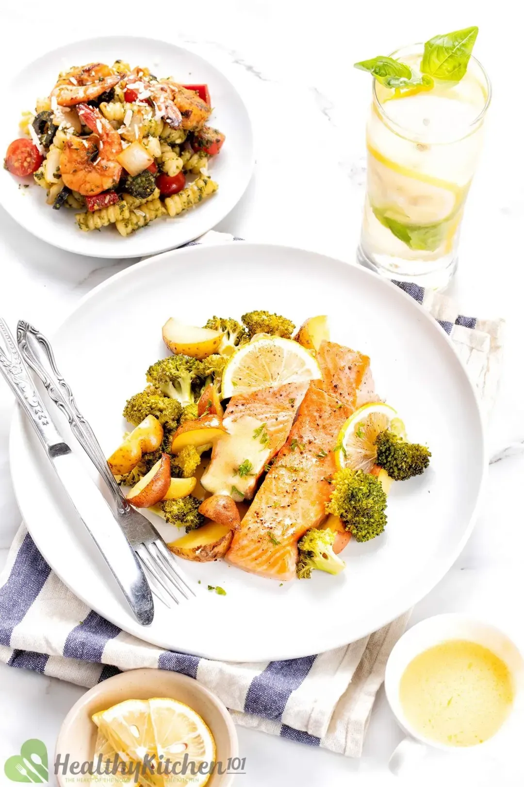 A serving plate of baked salmon, potato, and broccoli; on the side are a dish of pesto pasta salad, a tall glass of lemon water, a small bowl of buttery sauce, and lemon slices.