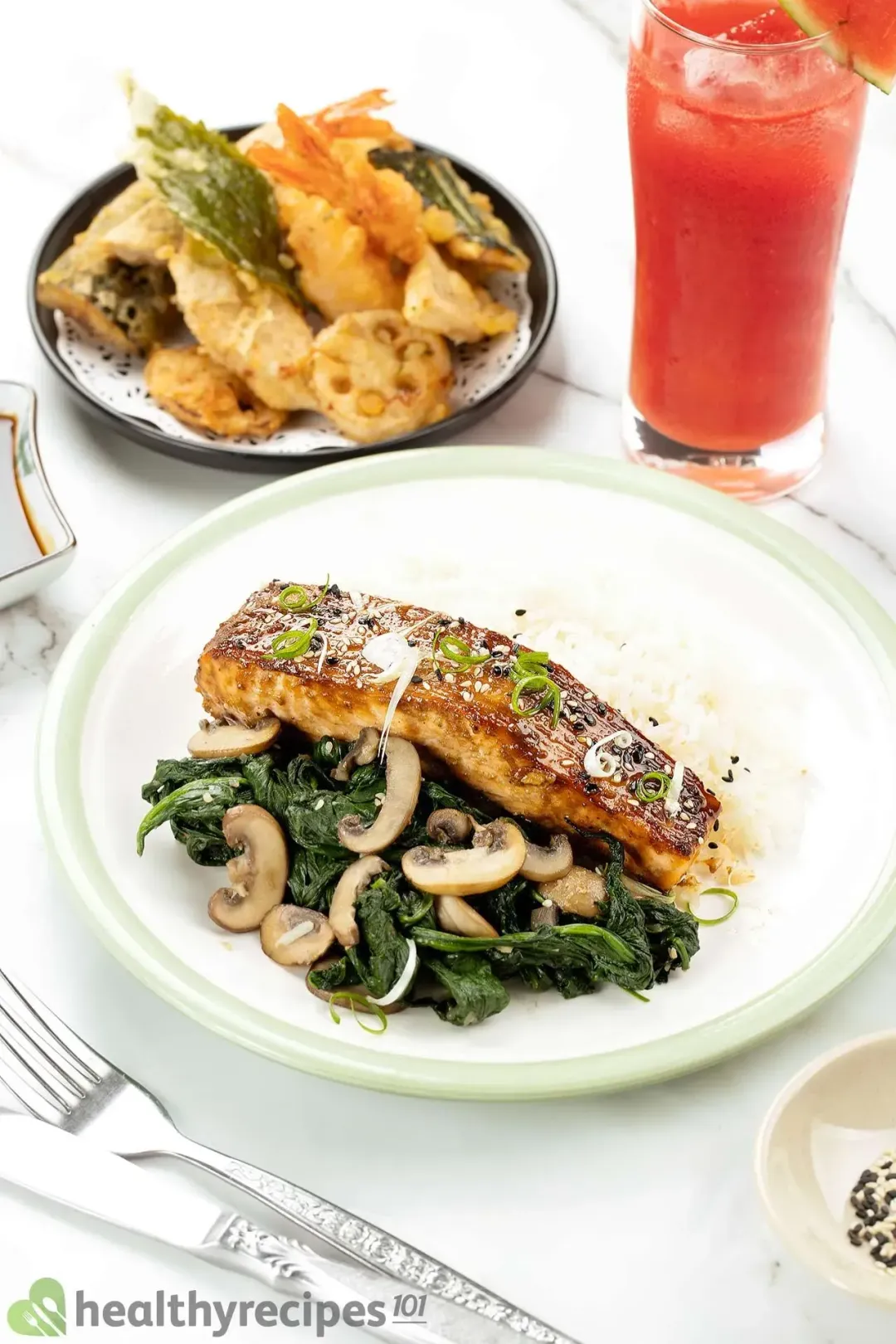 A plate containing a piece of miso-glazed salmon served alongside white rice, cooked mushrooms, and cooked spinach laid next to watermelon juice and tempura vegetables