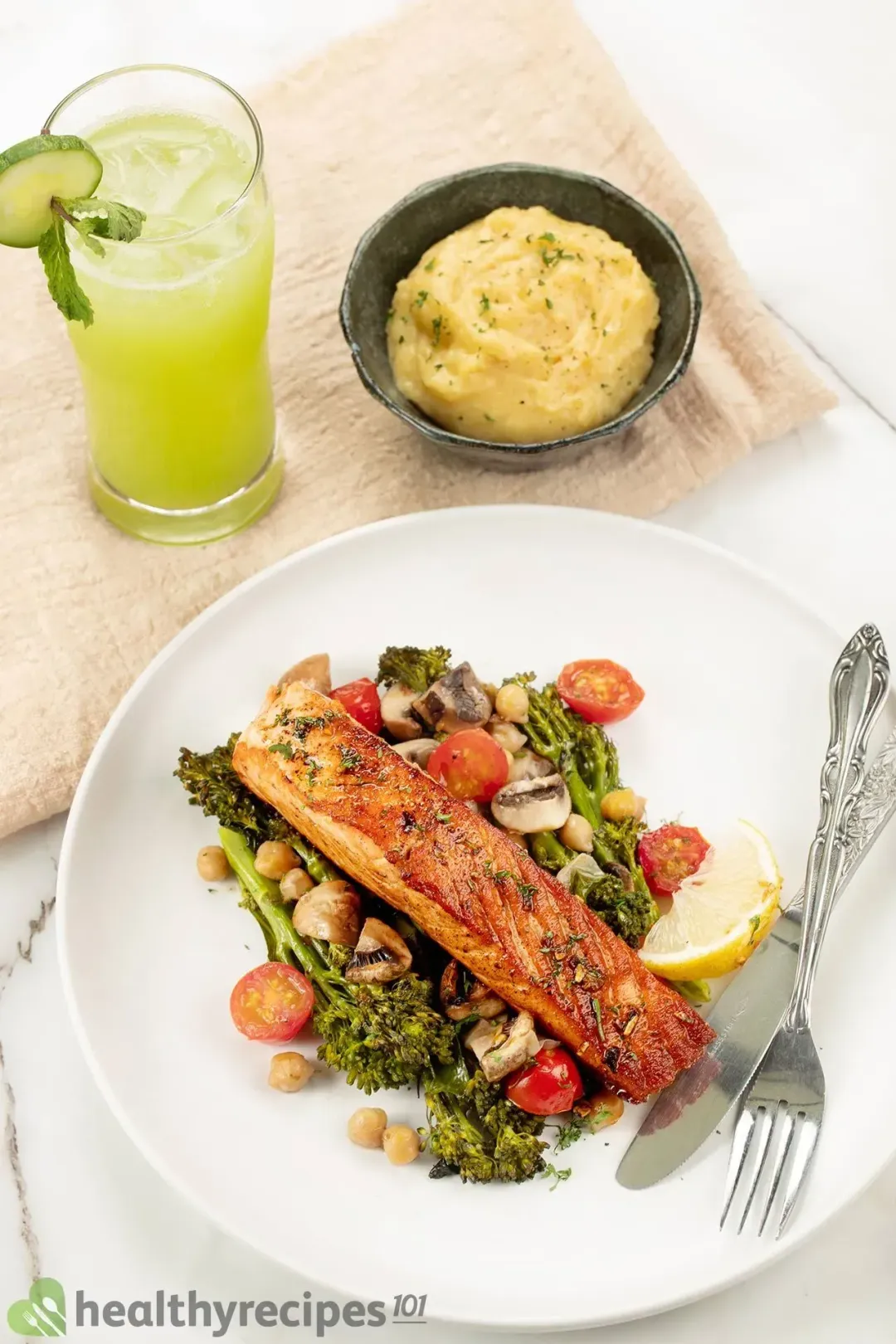 A plate of pan-seared salmon laid on a bed of vegetables next to a dinner knife and a fork. The dish is laid next to a green glass of cucumber juice and mashed potatoes.