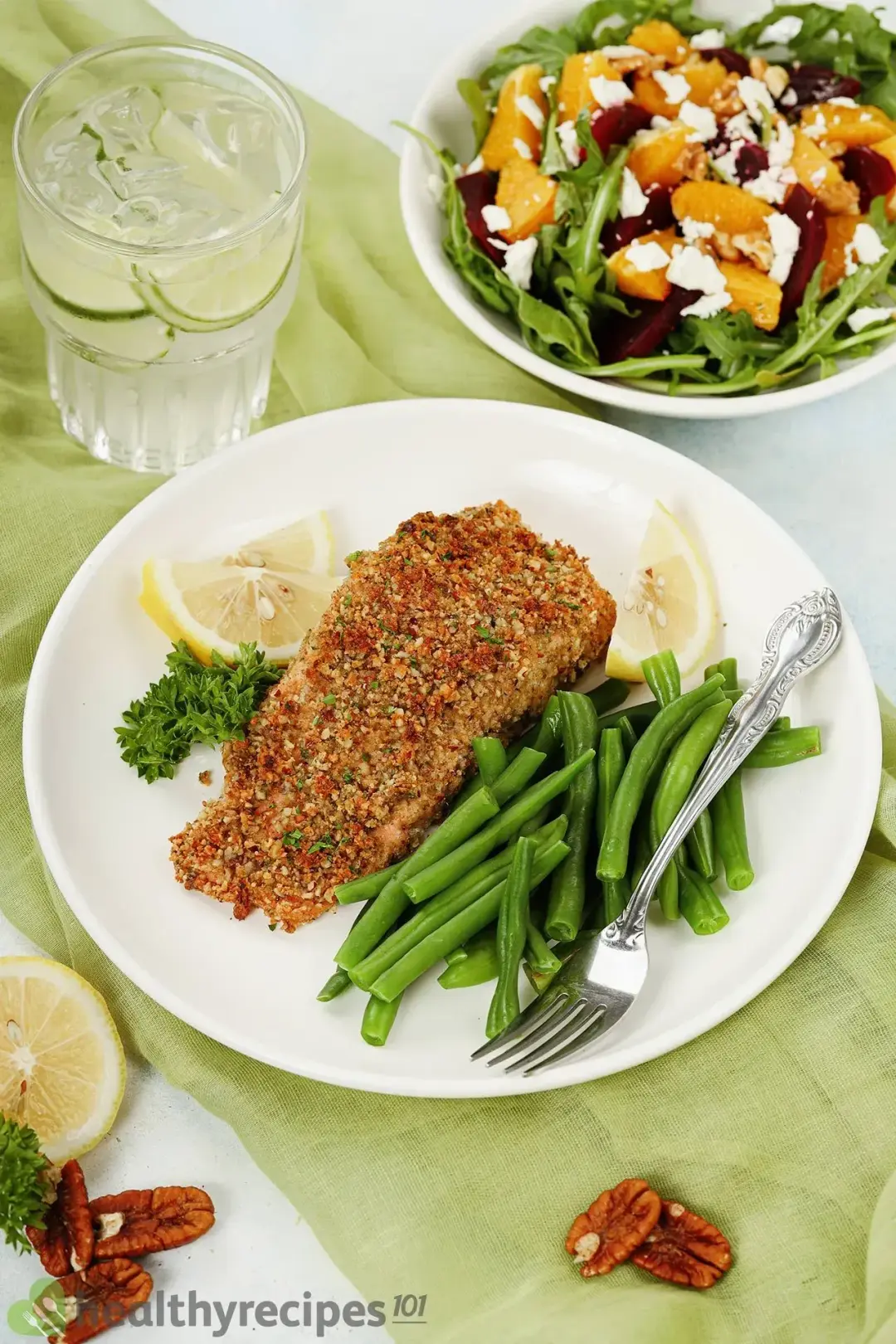 A big plate of one pecan-crusted salmon filet, cooked green beans, parsley, lemon slices, and a fork; the surroundings are a small plate of salad, a small cup of lime juice, a lemon slice, and some pecans.