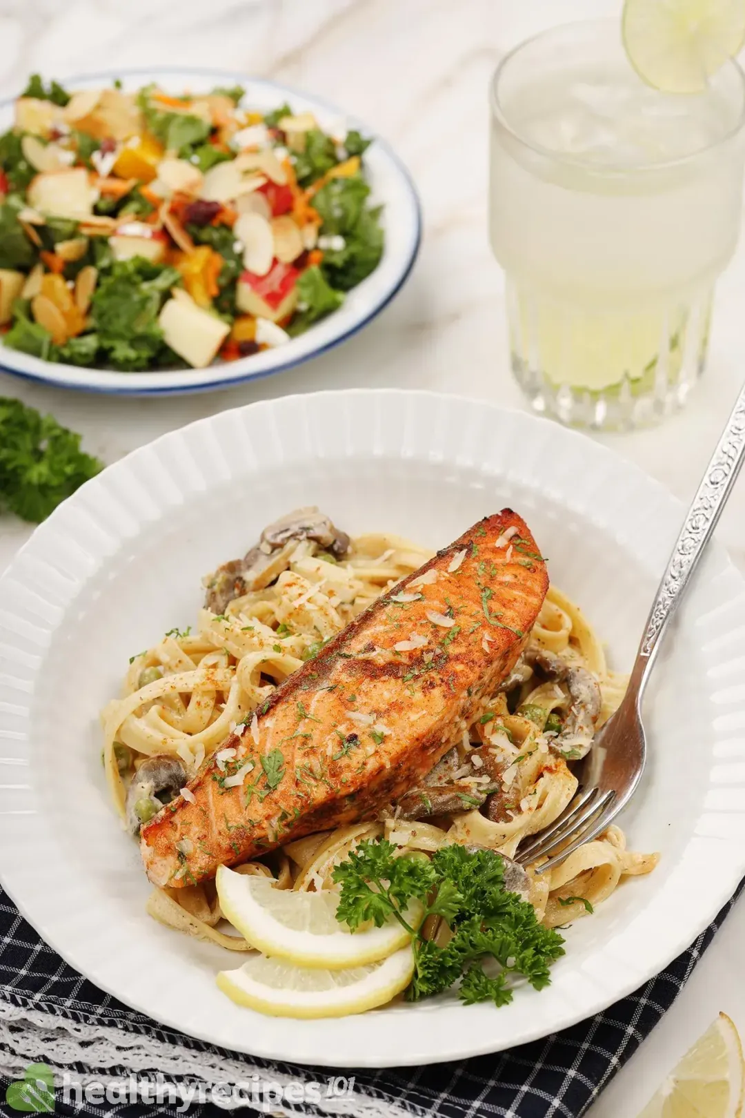 A plate of salmon al fresco, containing a bright orange cooked salmon fillet laid on a bed of fettuccine pasta, decorated by fresh parsley, lemon slices, and a fork