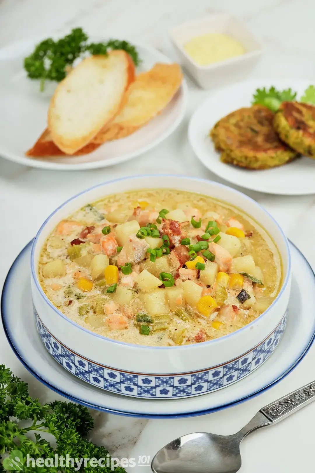 A bowl of salmon chowder with a plate of air-fried baguette slices and falafel on the background