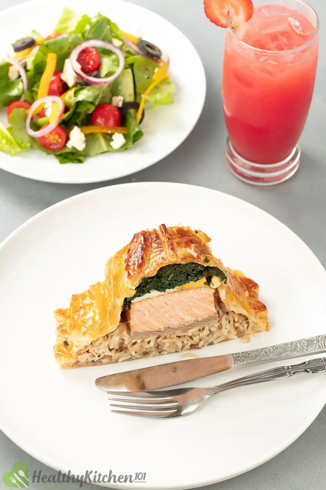 A slice of salmon coulibiac on a white plate with silver utensils, next to a Greek salad and iced watermelon juice