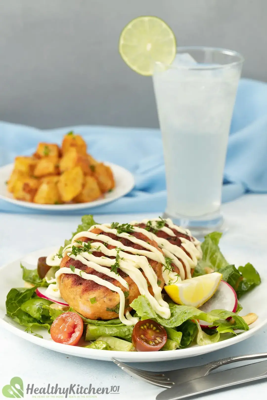 A picture of some salmon croquettes drizzled with the white sauce on a plate with fresh veggies and greens in a blurred background of a glass of lime juice and a plate of fried potatoes.