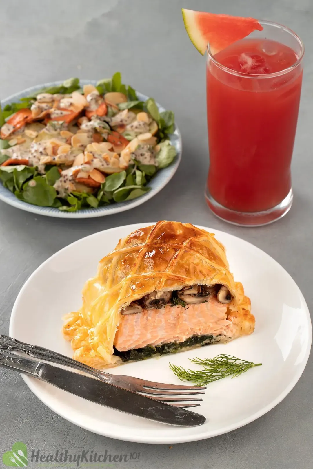 A plate of salmon en croute, a dinner knife, and a fork laid near a plate of mustard potato salad and a glass of watermelon juice