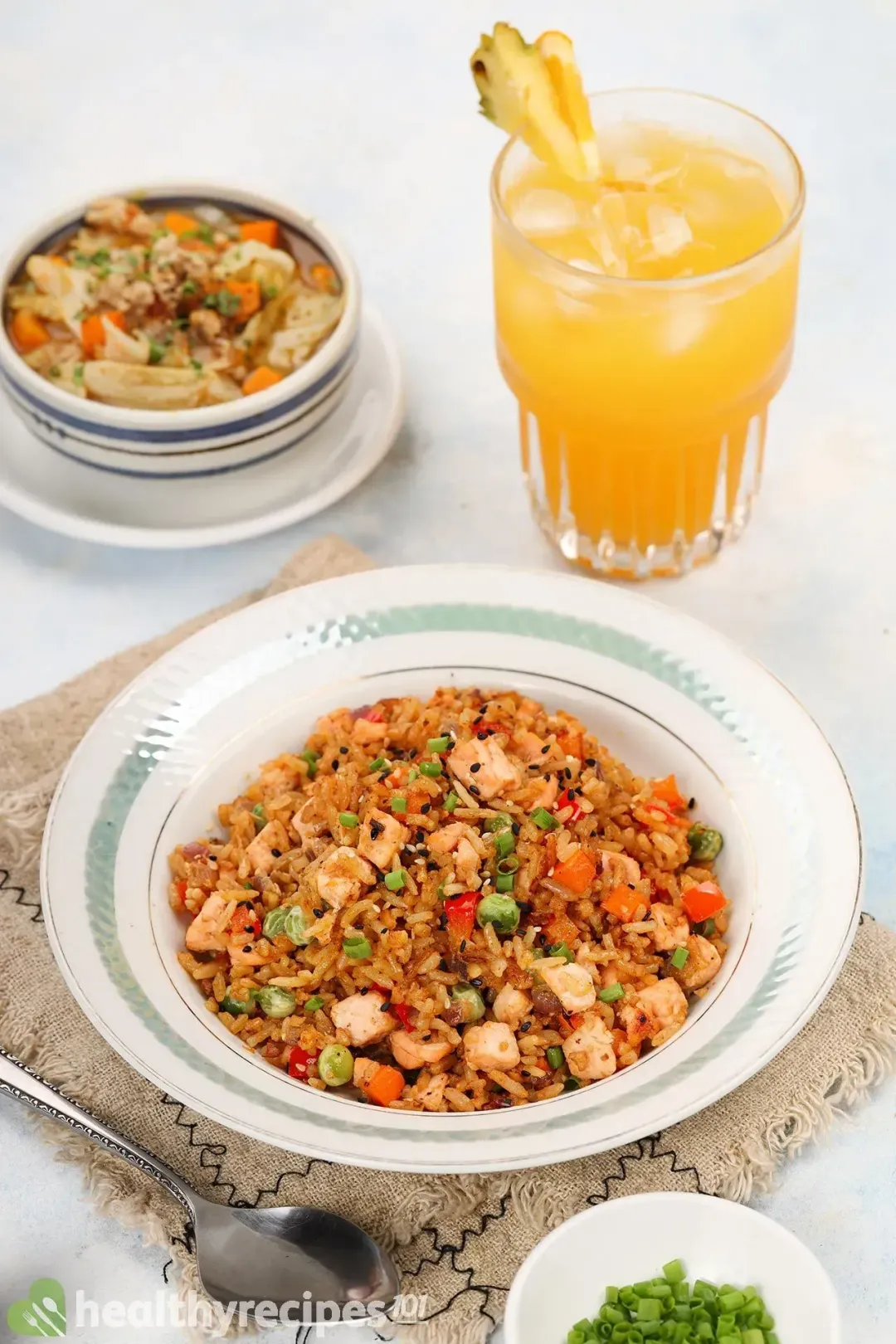 A white plate of salmon vegetable fried rice on a kitchen cloth, next to a spoon, a soup and an orange juice