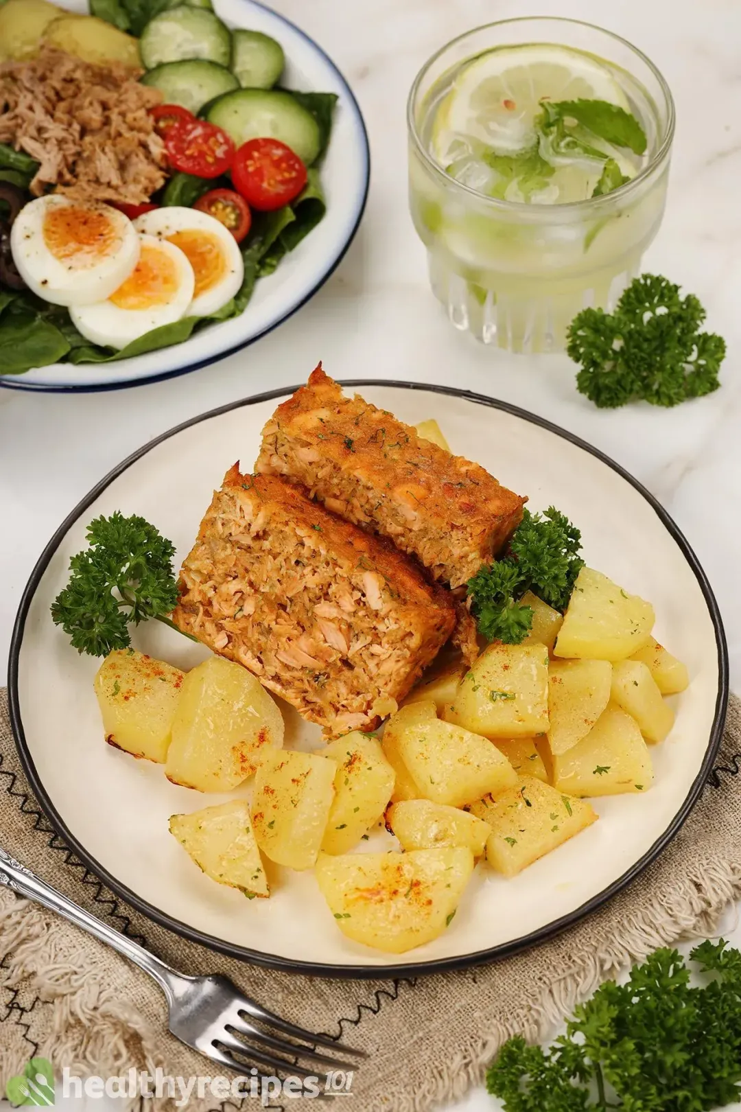 A plate containing salmon cakes and potato cubes decorated with fresh parsley, laid near a plate of salad and a glass of lime water