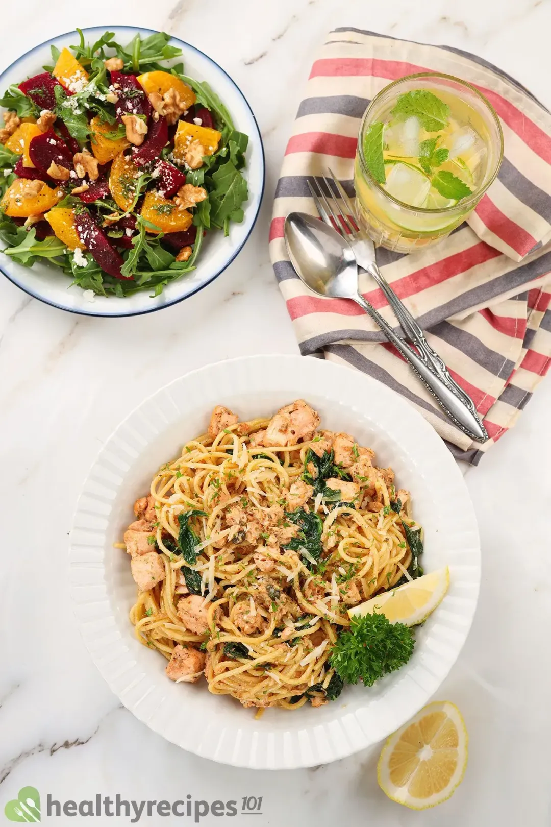 A high-angle shot of a plate of salmon pasta laid near a glass of lemon juice, a fork, a spoon, a striped tablecloth, a lemon wedge, and a beet orange salad