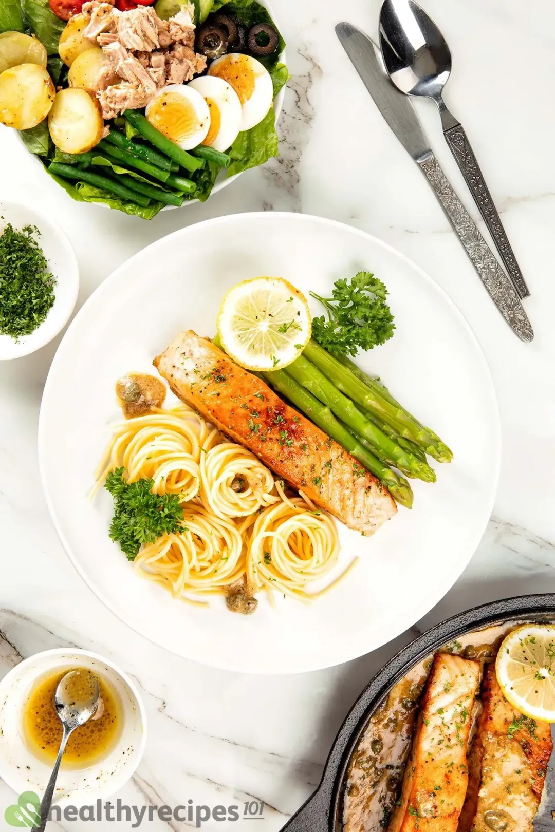 A plate of seared salmon served with cooked pasta and asparagus and topped with a lemon slice and parsley; the surroundings are a smaller plate of salad, a knife, a spoon, two small bowls of sauce and parsley, and two salmon filets in a skillet.