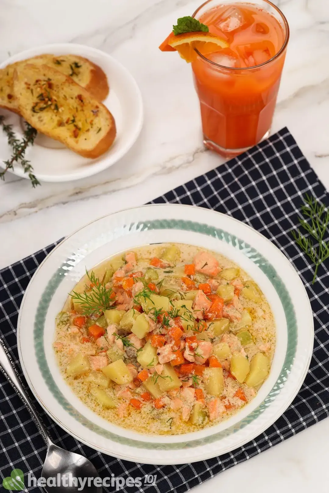 A deep plate of salmon soup laid on a blue tablecloth and next to a spoon, some toasted bread, and a glass of orange carrot juice