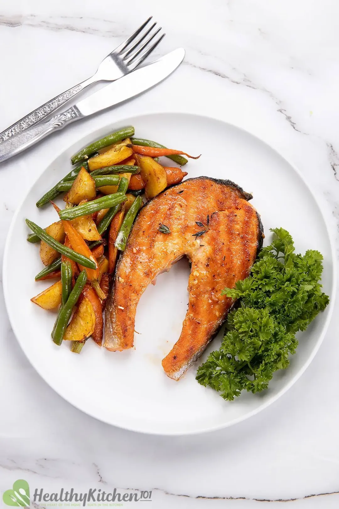 A portion of grilled salmon steak, roasted vegetables, and bundles of parsley on a white plate