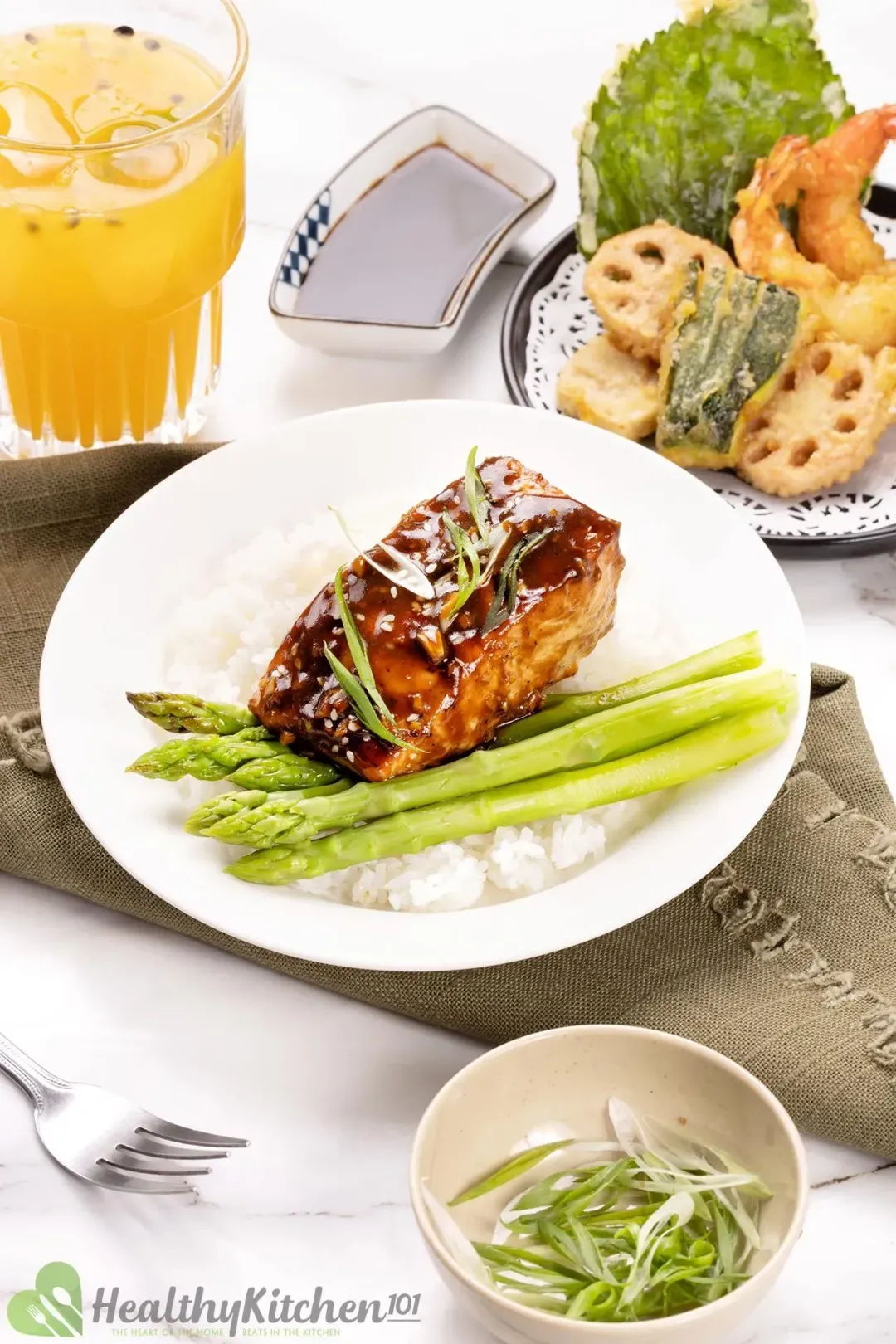 A serving plate of a baked teriyaki salmon filet placed on cooked white rice and asparagus; on the side are a fork, a smaller plate of tempura, a cup of passion fruit juice, two small bowls of teriyaki sauce and chopped scallion.