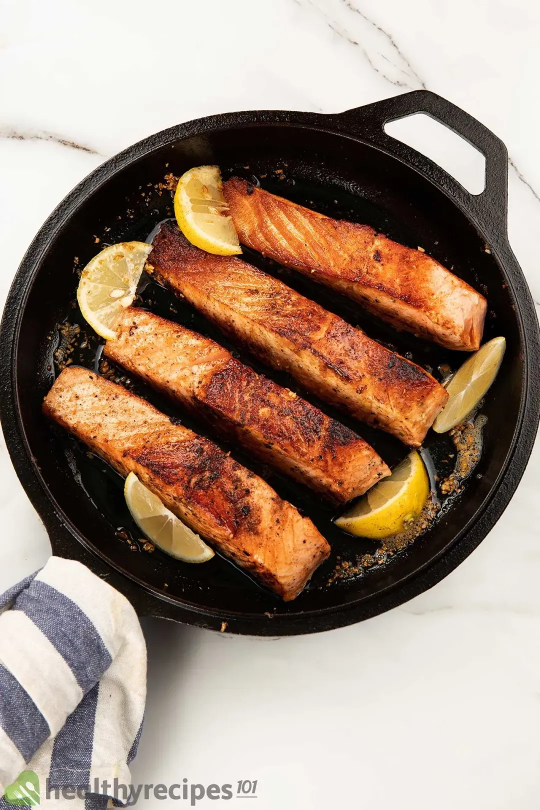 Four pan-seared salmon fillets with brown edges laid on a skillet with a few slices of lemon