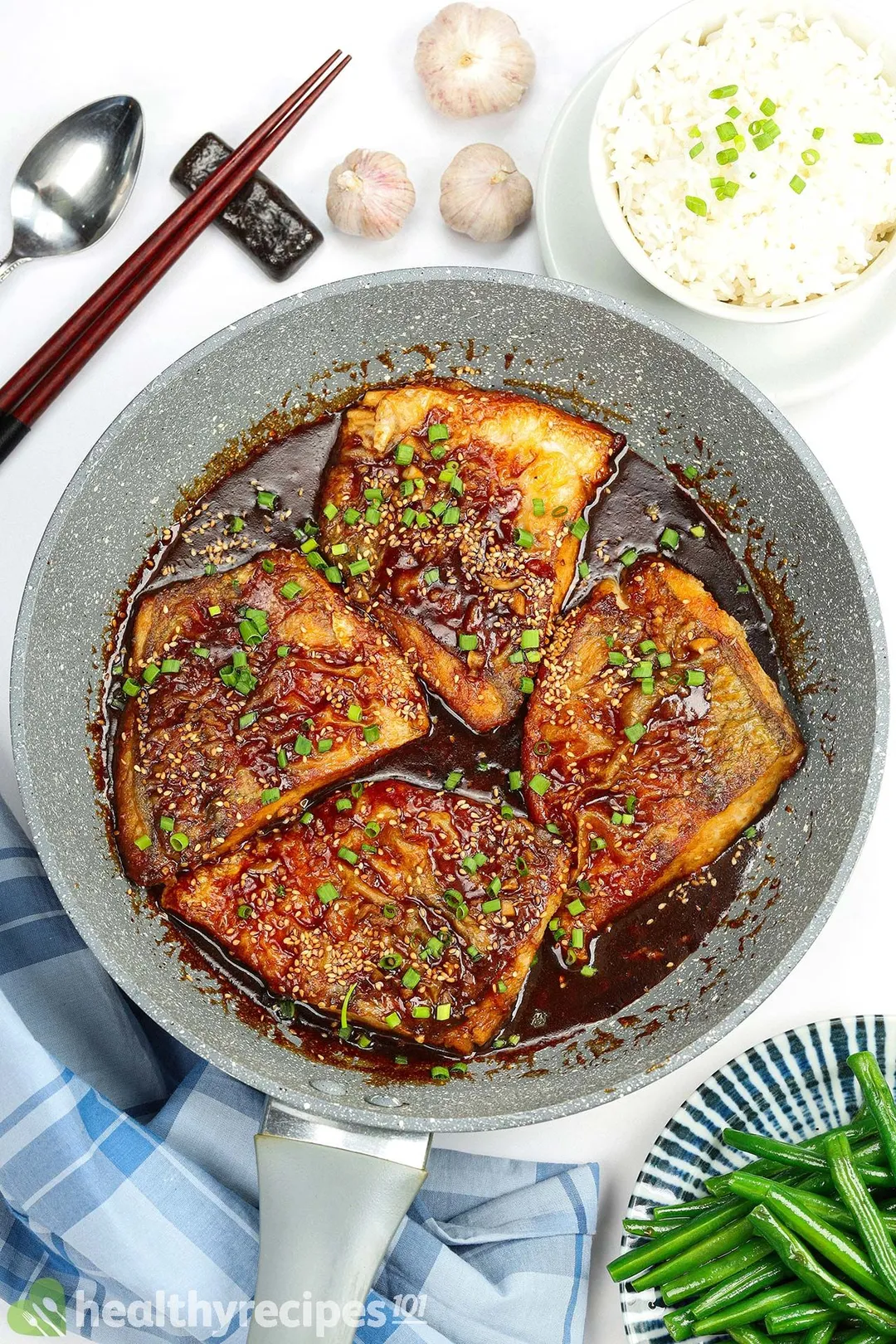 a non-stick skillet of cooked sea bass surrounded by rice, green been, garlic, spoon and a pair of chopstick