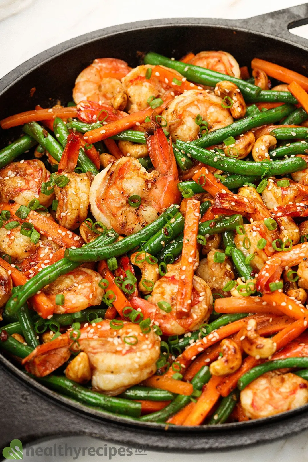 A dish full of cooked shrimp, green beans, and carrot strips, surrounded by a soup bowl, a rice bowl, a soy sauce bowl, a watermelon drink, and split cashews