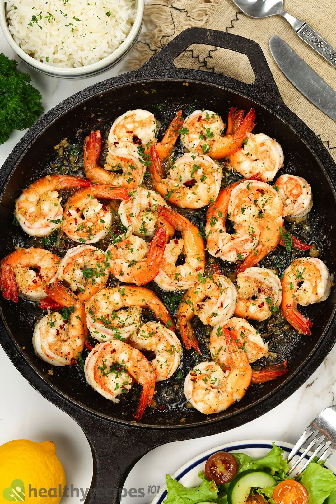 a cast iron skillet of cooked shrimp with chopped parsley on top decorated by lemon, salad dish, spoon and knife and a bowl of cooked rice 