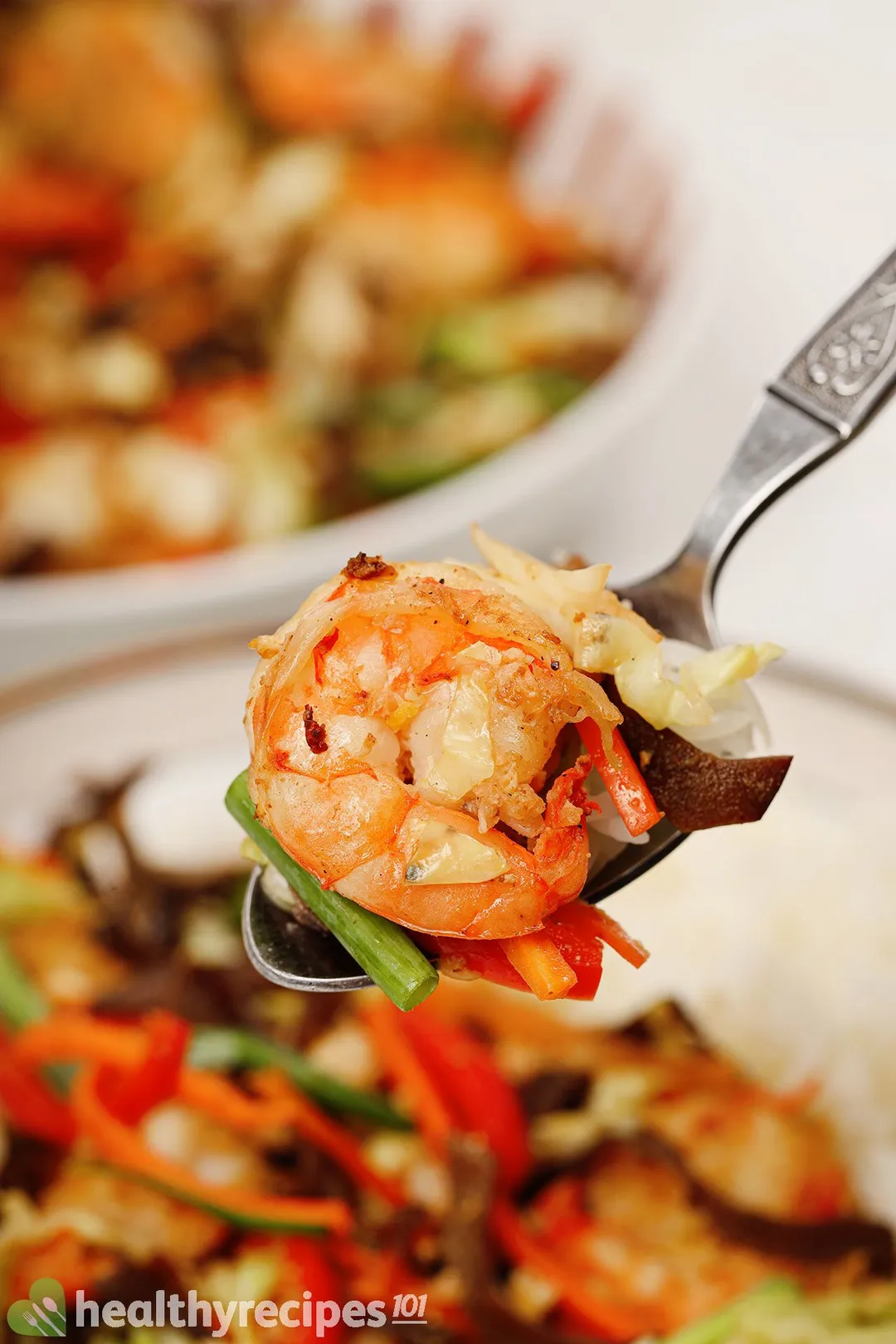 A spoon holding a piece of cooked shrimp, with colorful plates of vegetable saute in the background