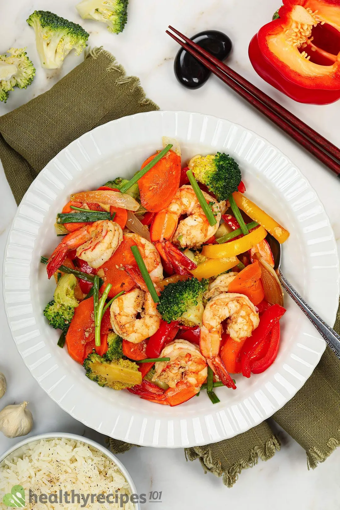 Pan-fried shrimp and vegetables served in a plate next to a bowl of rice, a pair of chopsticks, half a bell pepper, garlic bulbs, and some broccoli florets.