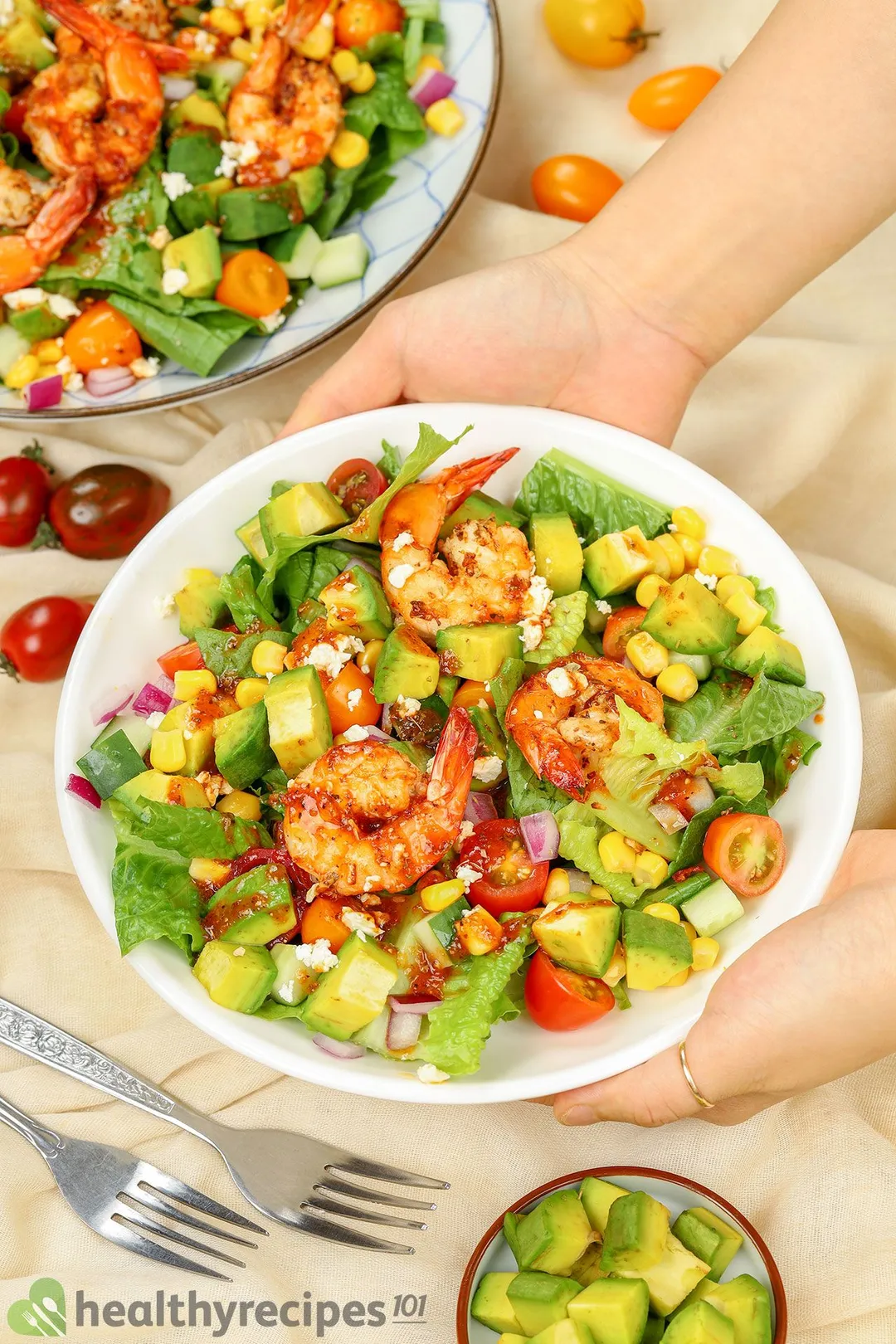 two hands holding a plate of cooked shrimp and avocado cubes salad decorated by a small plate of avocado cubes and some cherry tomatoes and fork