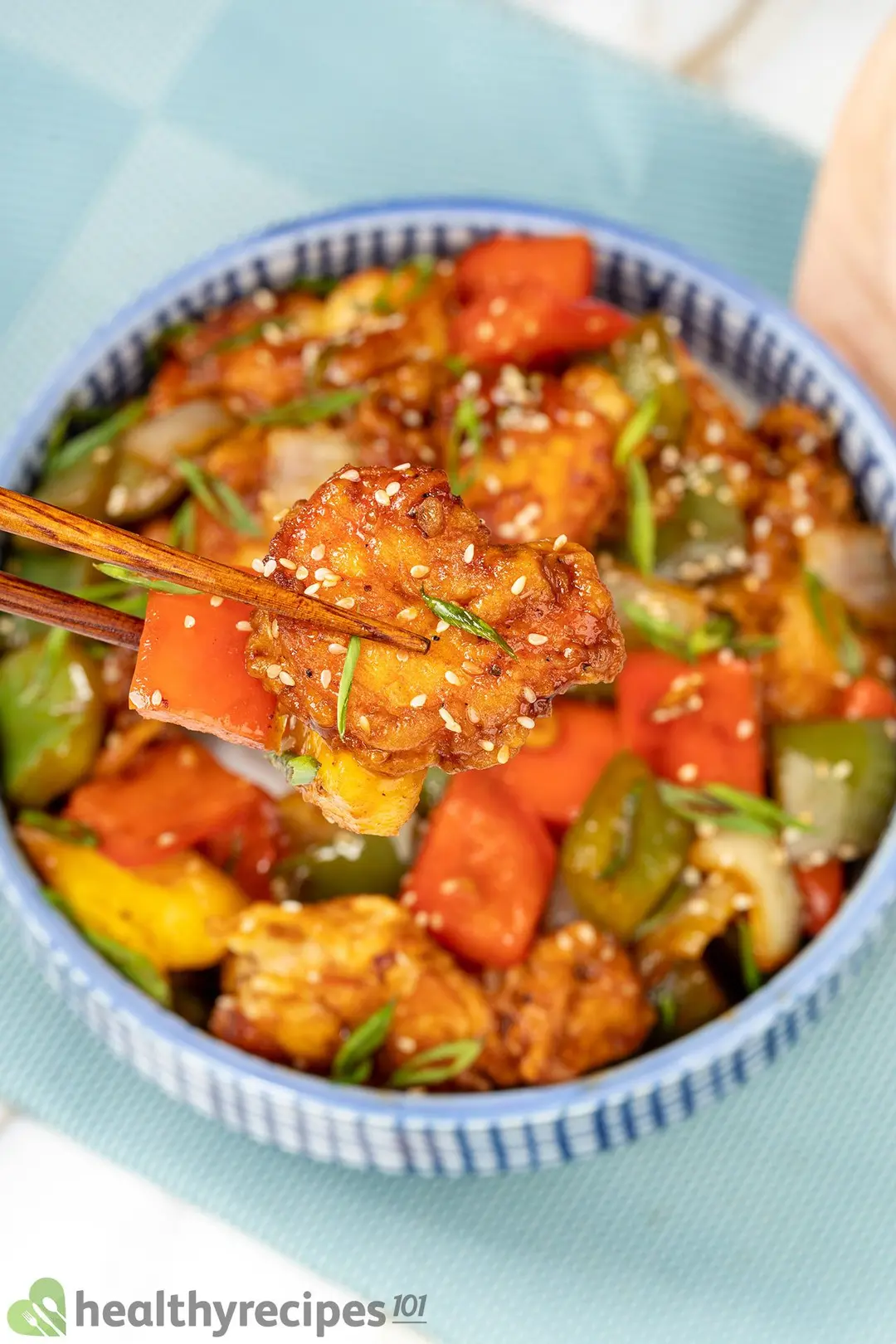 A blue-checkered bowl of battered shrimp sauteed with colorful vegetables, put next to a wooden pair of chopsticks
