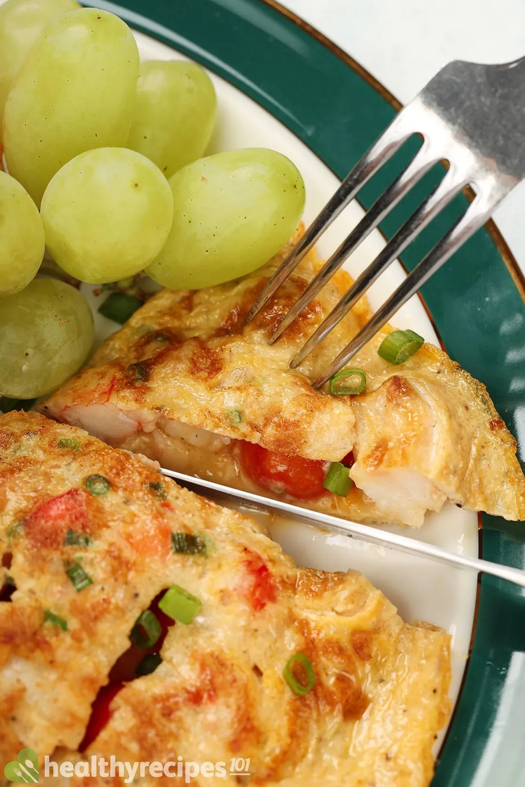 A fork piercing into a slice of shrimp omelette placed next to green grapes.