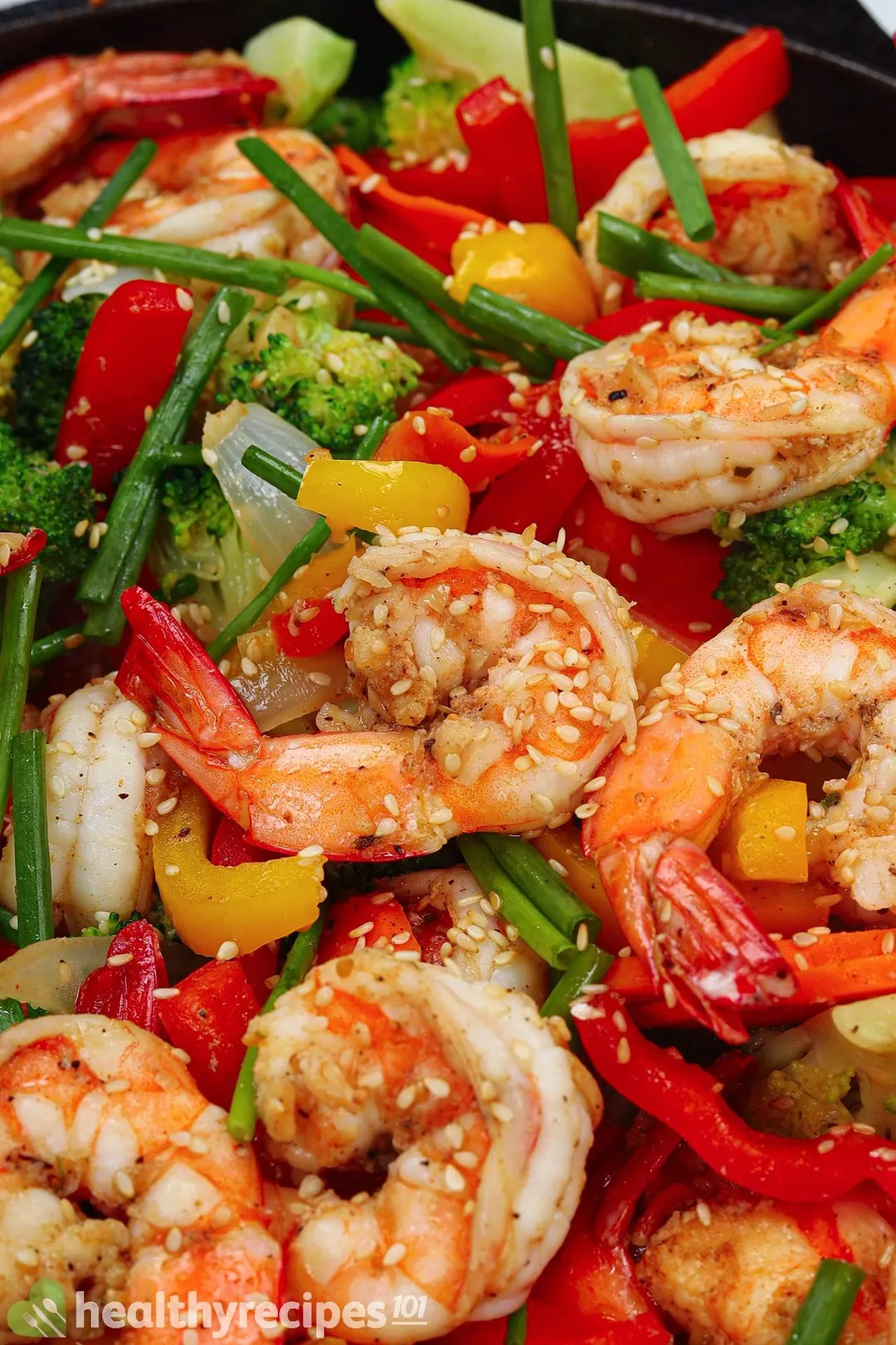 Shrimp sautéed with broccoli and bell pepper in a black skillet topped with scallion and sesame seeds.