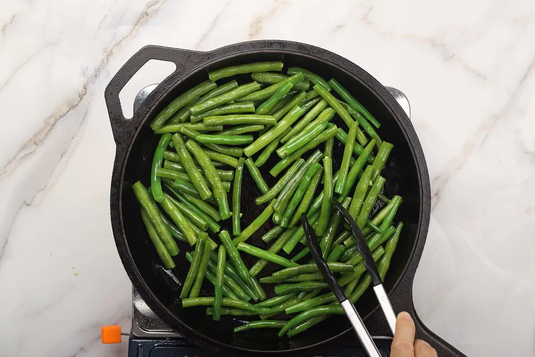 Green beans stirred in a cast iron skillet with a pair of tongs