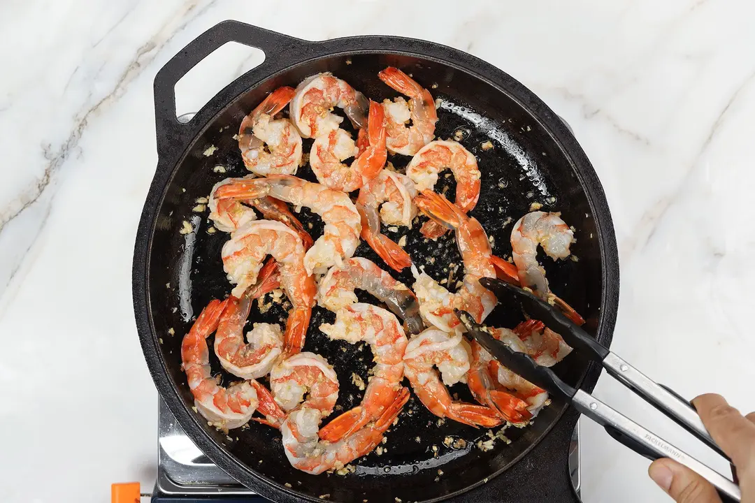 Shrimp being cooked with a pair of tongs inside a black cast-iron skillet