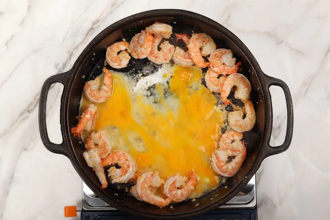A cast iron skillet with cooked shrimp on the rind, beaten egg about to set in the middle