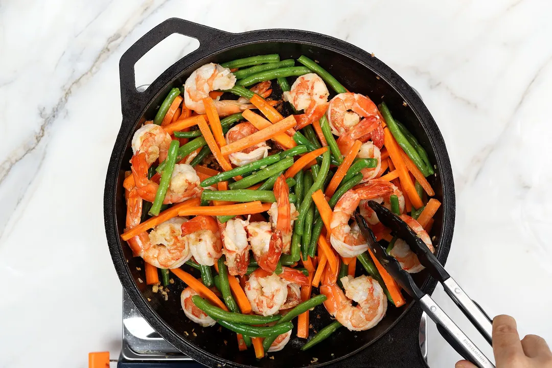 Shrimp, green beans, and thin carrot fingers stirred with a pair of tongs inside a black cast-iron skillet