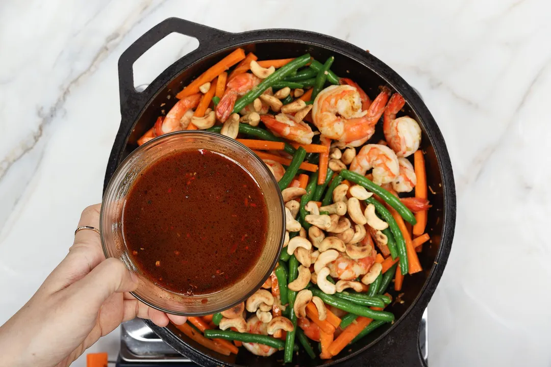 A hand holding a small bowl of brown sauce hovering over a cast-iron skillet of cooked shrimp, green beans, carrots, and cashews