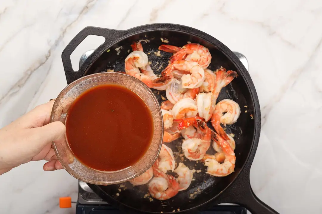 A hand holding a bowl of red sauce over a shrimp skillet