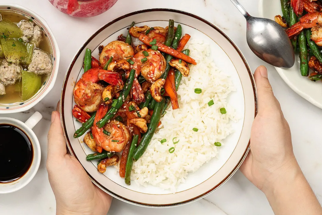 Two hands putting a dish of cooked white rice, cooked shrimp, green beans, carrots, and cashews next to a soup bowl, soy sauce bowl, and a dish of food with a metal spoon