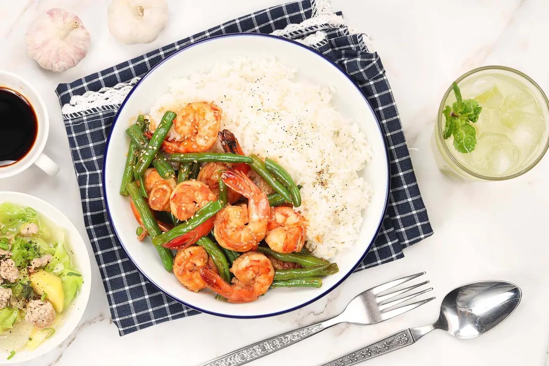 A meal consisting of a bowl of white rice, cooked shrimp and green beans placed on top of a blue-checkered cloth, put next to utensils, an iced drink, and a soup bowl