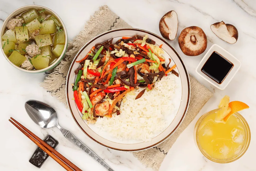 Rice served with a colorful vegetable saute in a plate put next to a soup bowl and an orange iced drink