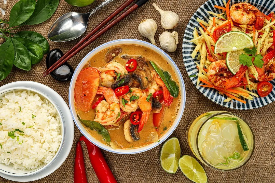 a bowl of tom yum soup next to a bowl of cooked rice, a plate of salad and a glass of juice