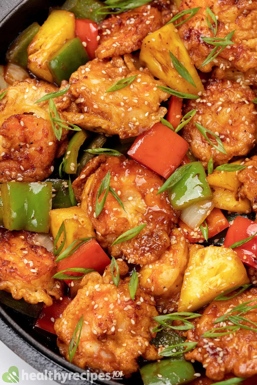 A close-up shot of battered-shrimp cooked with chunks of red peppers, green peppers, and pineapples, garnished with thinly sliced scallion and white sesame seeds