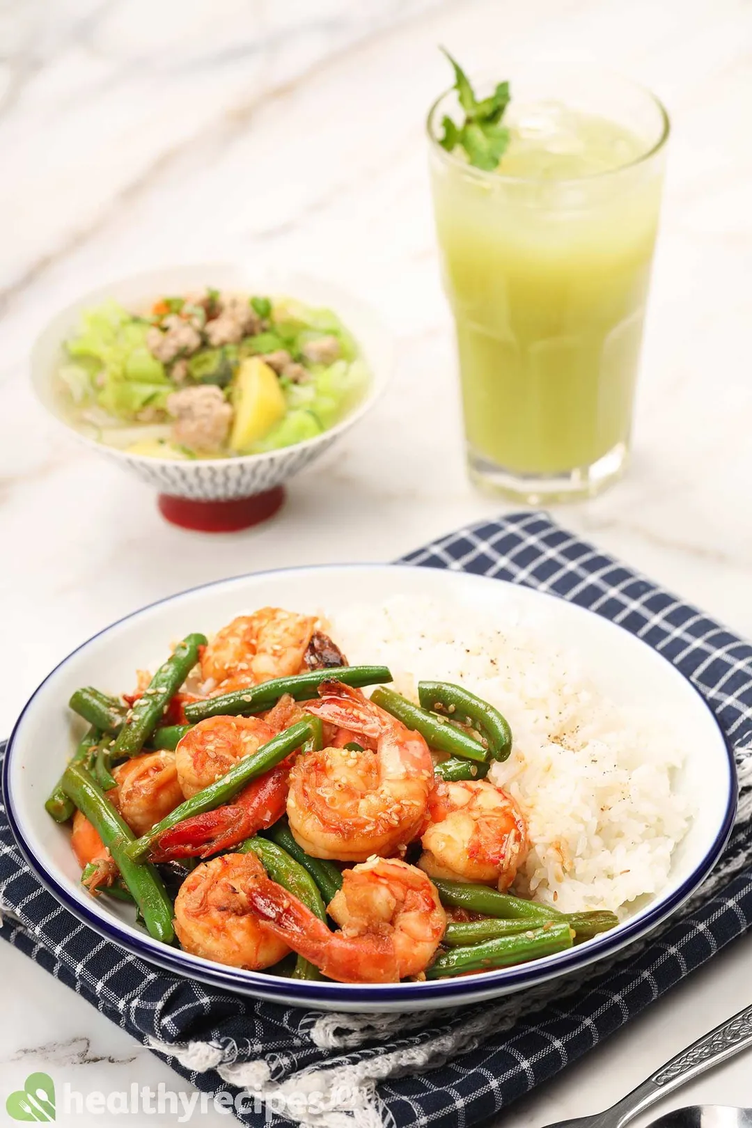 A white dish filled with rice, shrimp, and green beans put on a blue checkered tablecloth, with a bowl of soup and a glass of green juice in the background