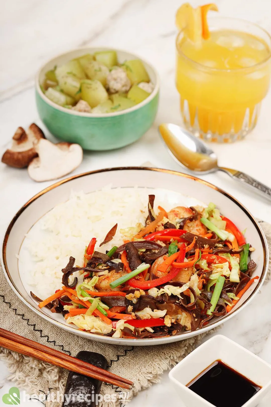 A plate of rice and colorful shrimp vegetables saute, with a soup bowl and iced orange drink in the background