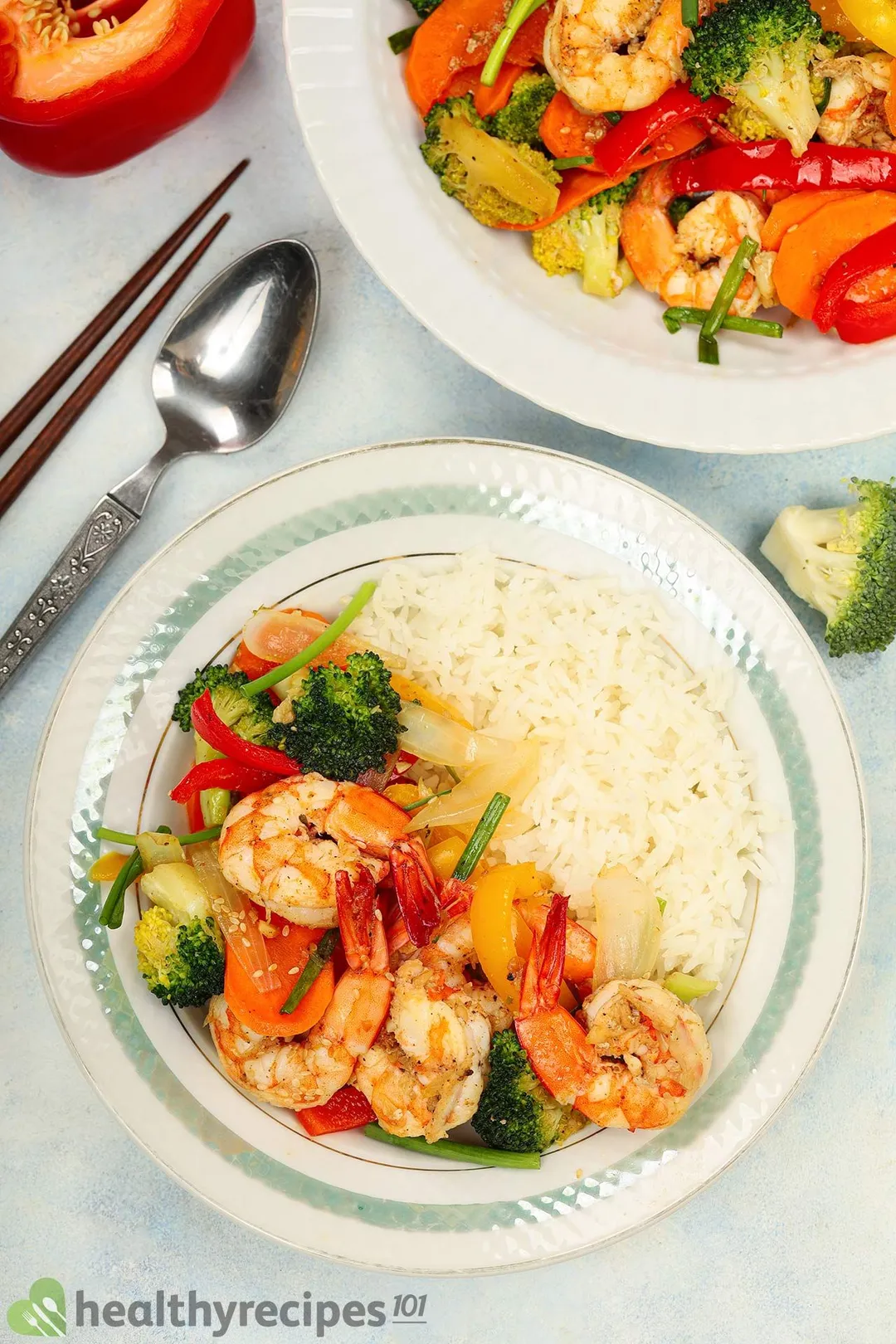Pan-fried shrimp and vegetables served with rice on plates next to a spoon, a pair of chopsticks, half a bell pepper, and a broccoli floret.