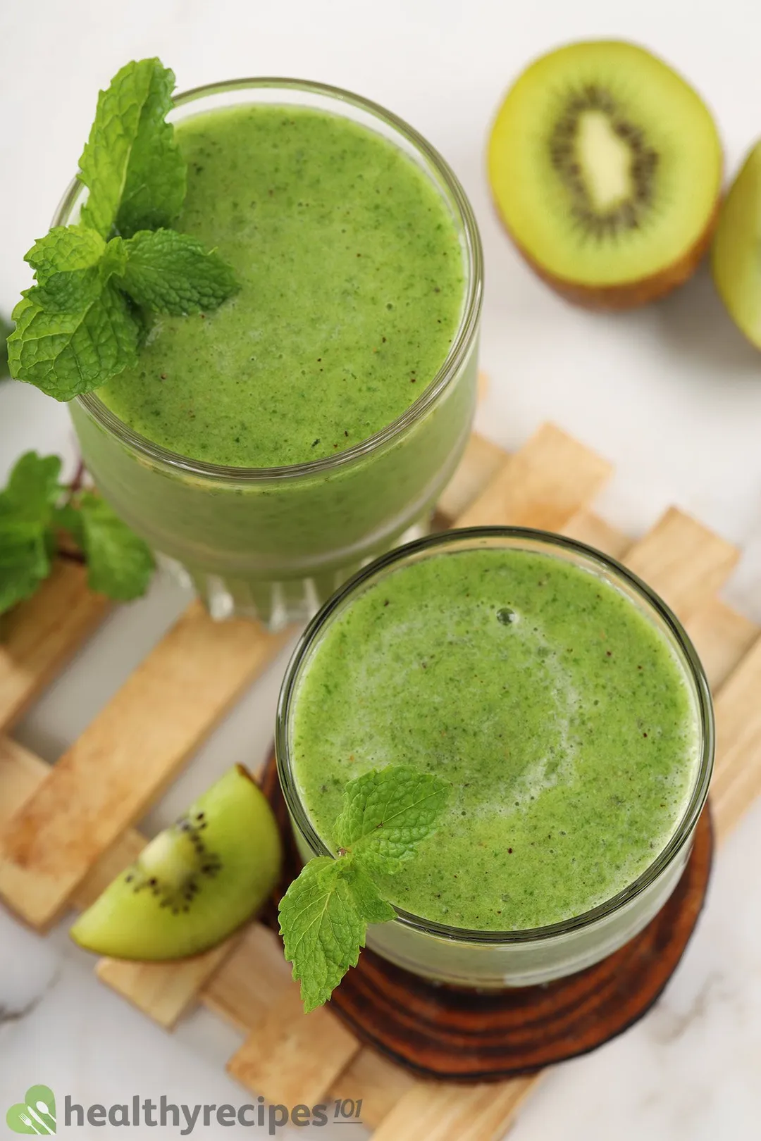 A high-angle shot of two glasses of apple kiwi kale smoothie laid on a wooden plank and surrounded by sliced kiwis.