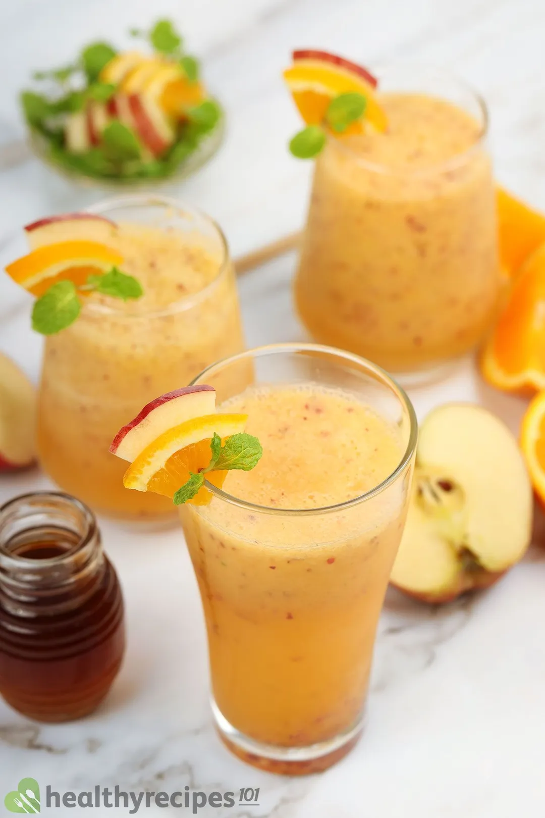 Three glasses of apple orange smoothie laid near a small jar of honey, half an apple, and some orange wedges.