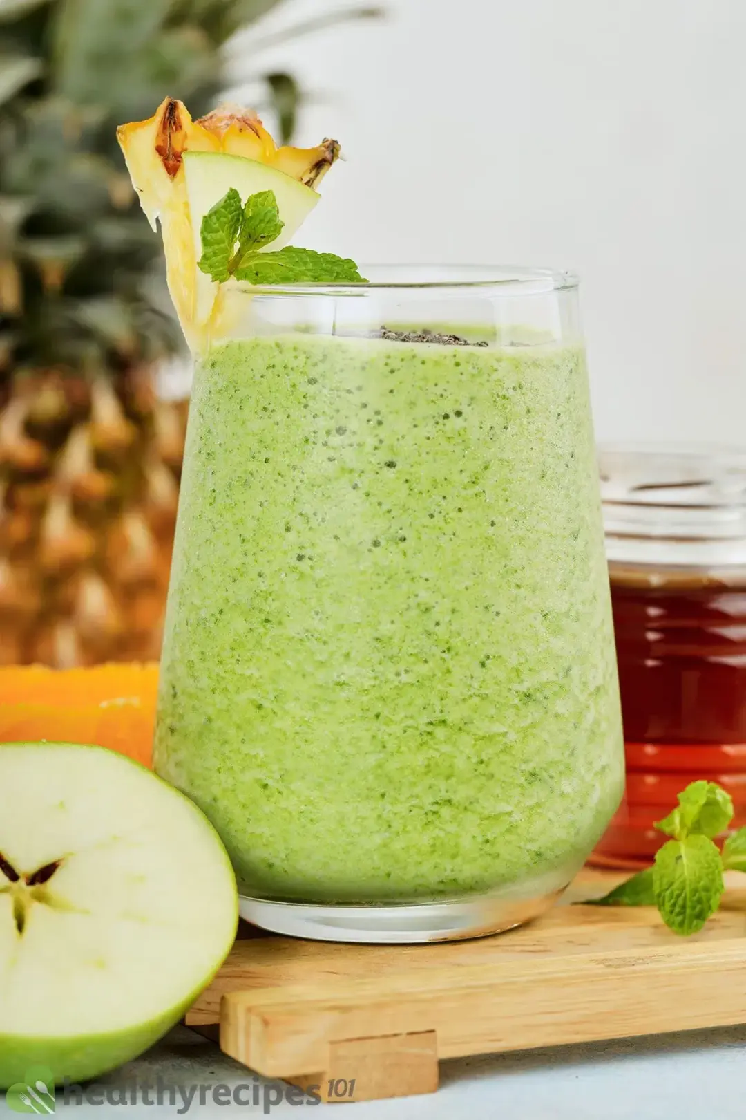 A glass of green apple smoothie surrounded by half a green apple and a small jar of honey