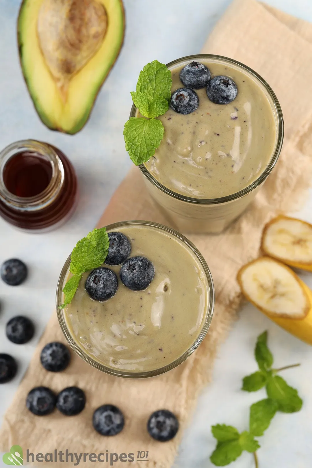 A high-angle shot of two glasses of avocado blueberry smoothie, surrounded by half an avocado, blueberries, sliced bananas, mint leaves, and a jar of honey.
