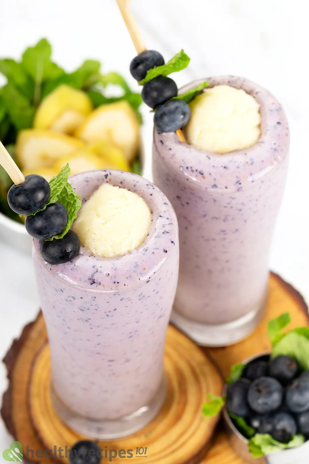 Two tall glasses of blueberry banana smoothie placed on wooden boards and surrounded by blueberries, sliced bananas, and mint leaves.