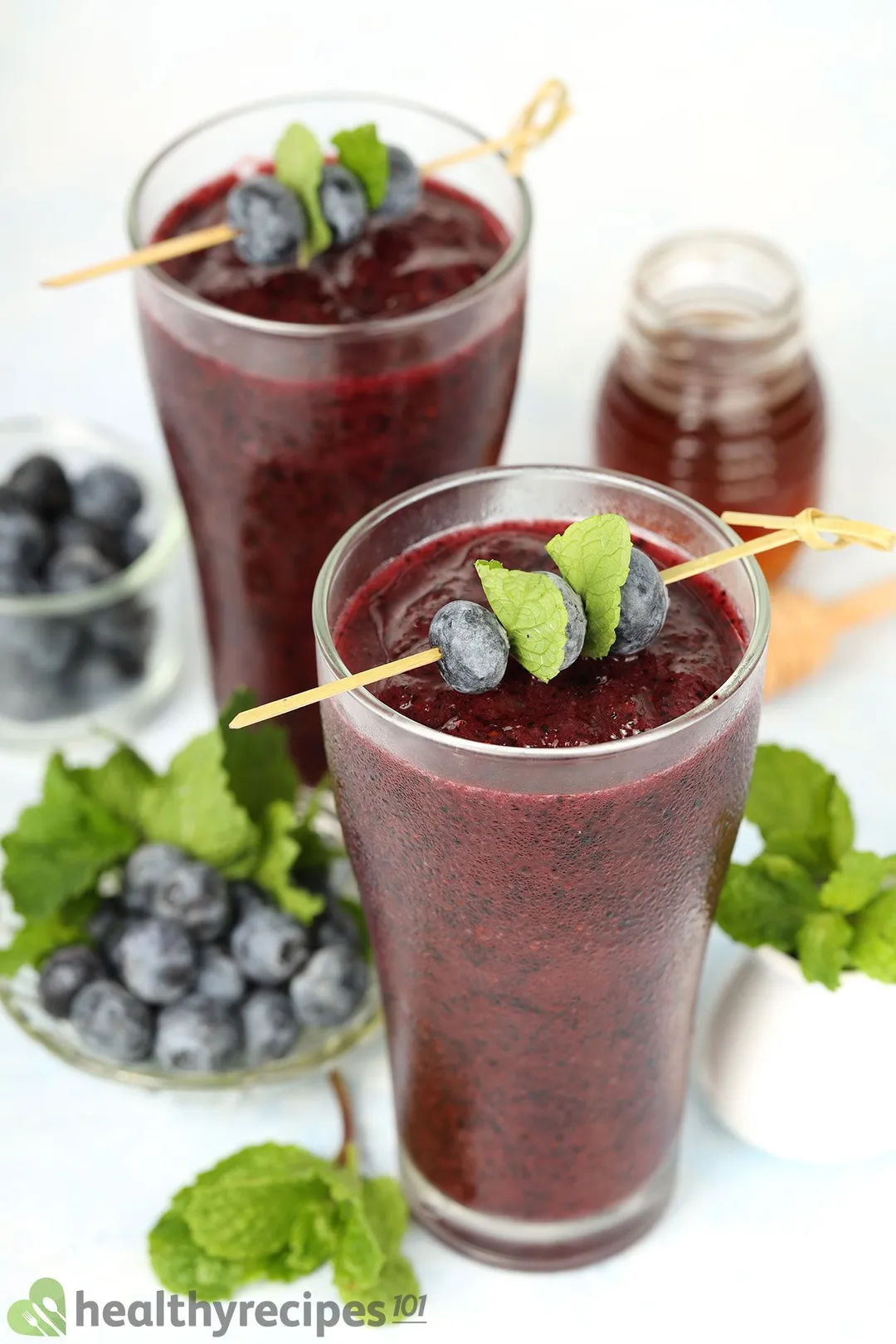 Two glasses of blueberry smoothie garnished with blueberry skewers and placed near a small plate of blueberries and a small jar of honey.