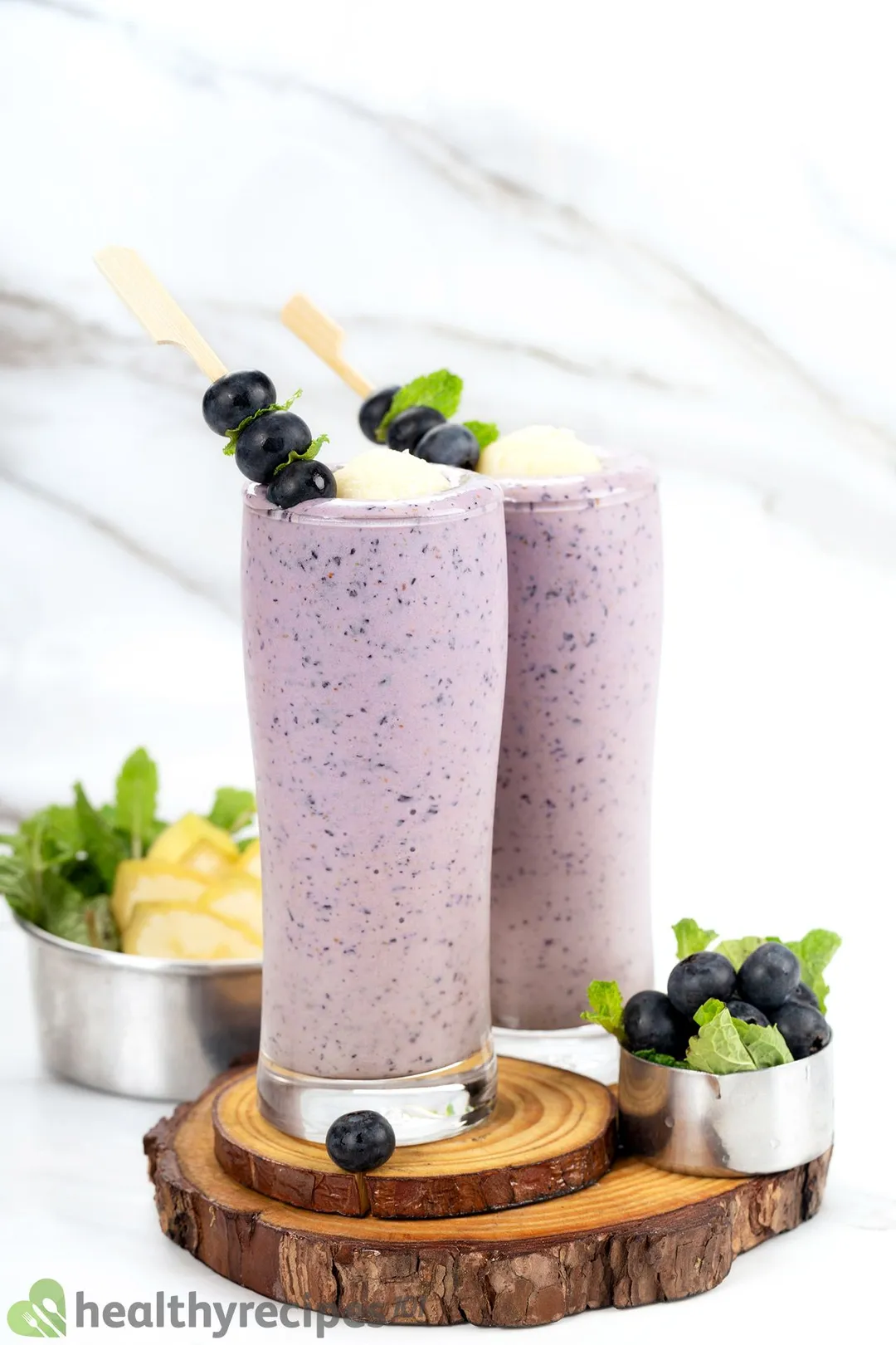 Two tall blueberry banana smoothie glasses placed on wooden boards with blueberries nearby and a small bowl filled with bananas and mint leaves in the background.